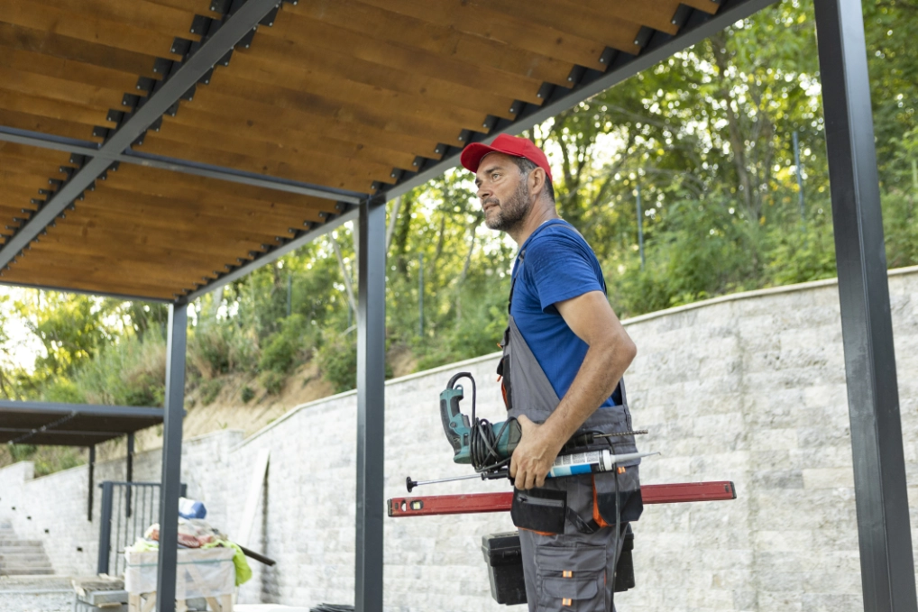 Monteur in grauem Overall steht mit Werkzeug unter einem Lamellendach